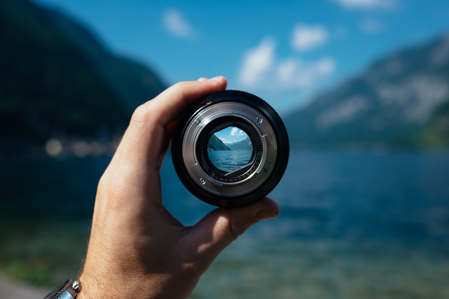 image of a hand holding a camera's lens, in front of a lake. Through the lens, the lake looks clear with the scene outside of the lens blurry