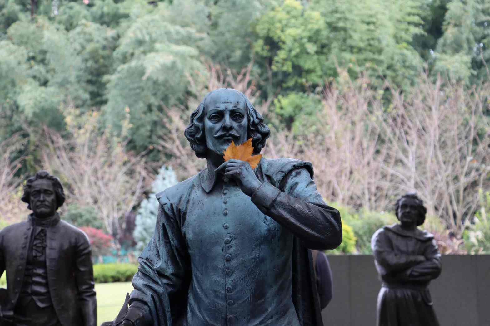 A statue of William Shakespear, with his hand up by his chin. There's a maple leaf sitting in the gap between his hand and chin, making it seem as if he's holding it.